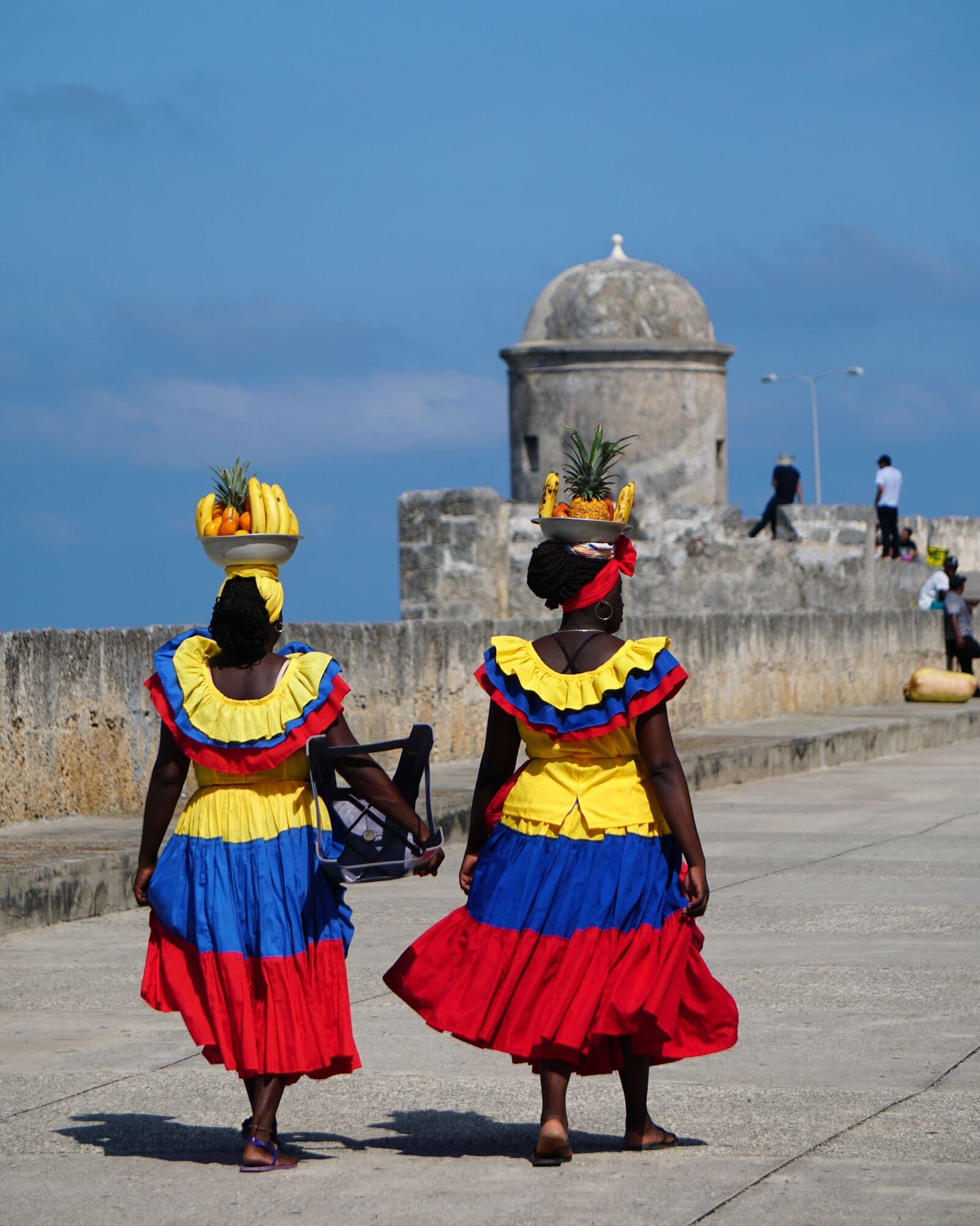 Two palenqueras in the yellow, blue and red dresses of Colombia walking along the city walls
