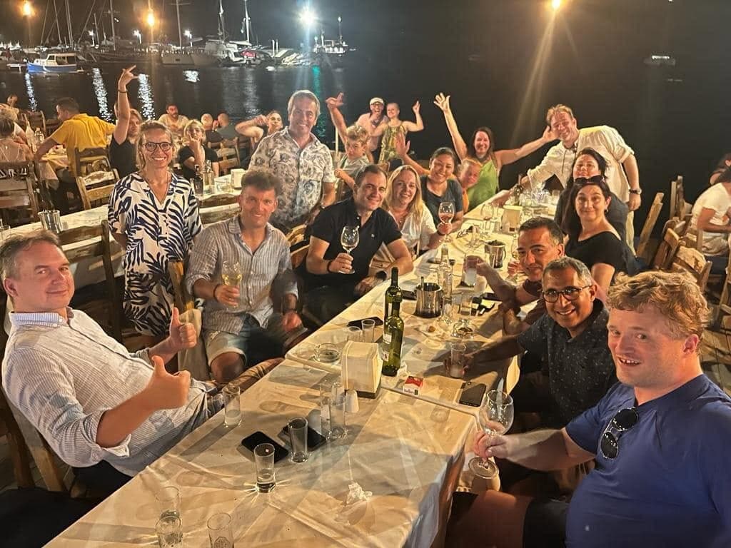 A long, loud dinner table by a harbour at night, glasses up